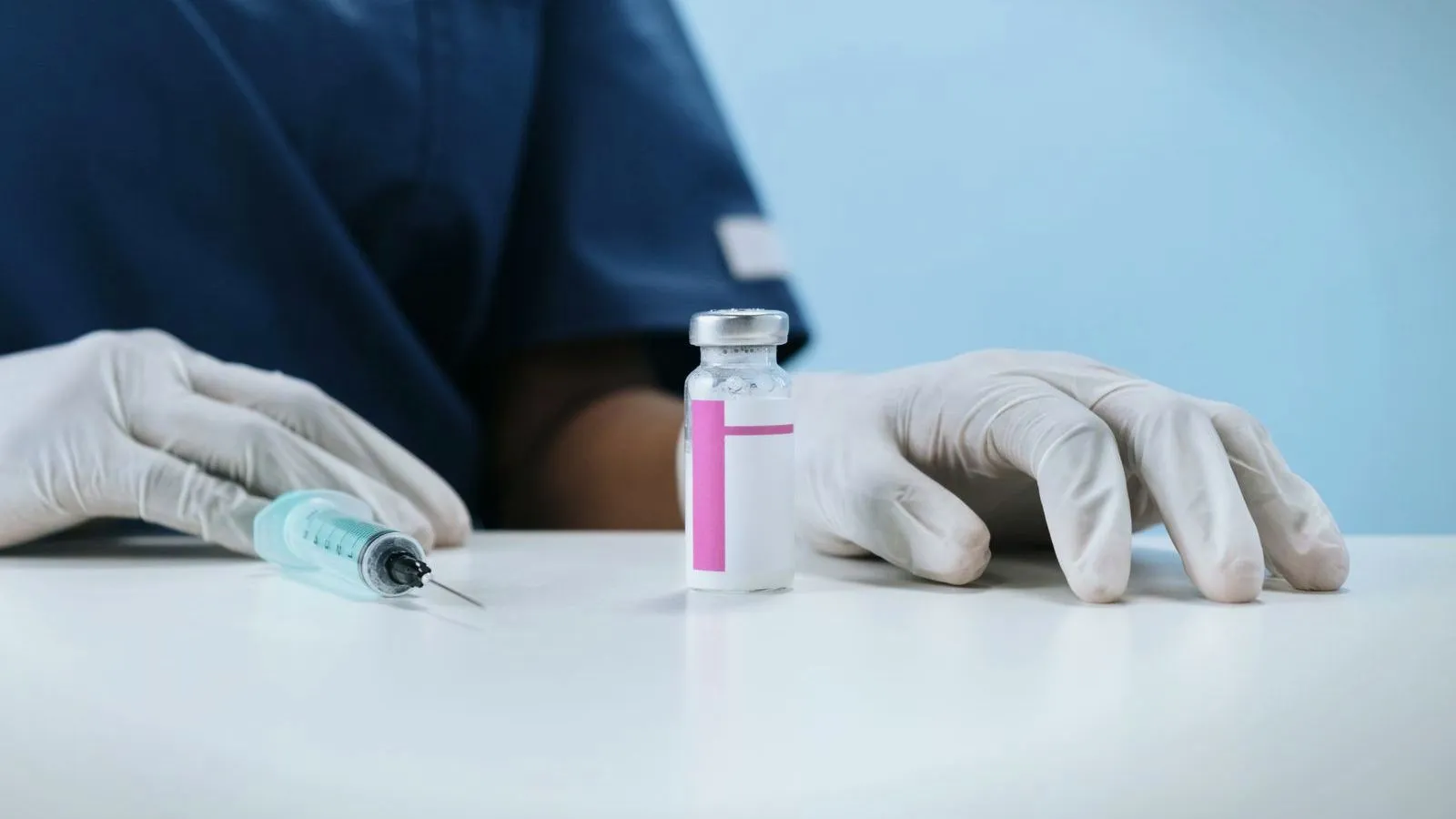 Gloved hands rest on a white table beside a vaccine vial and a filled syringe, with a person in dark blue scrubs behind.