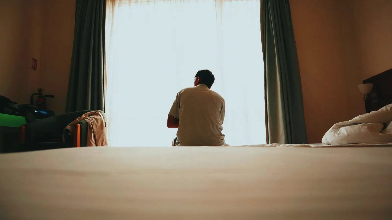 A man seen from behind sits on the edge of a bed facing a bright, curtained window in a dim bedroom.