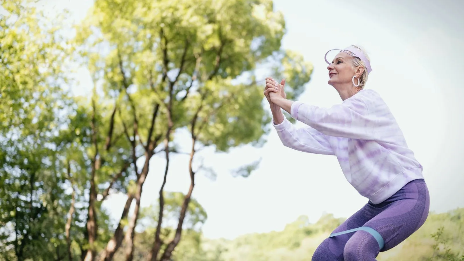 A woman in her fifties exercising outdoors, doing a squat with a resistance band