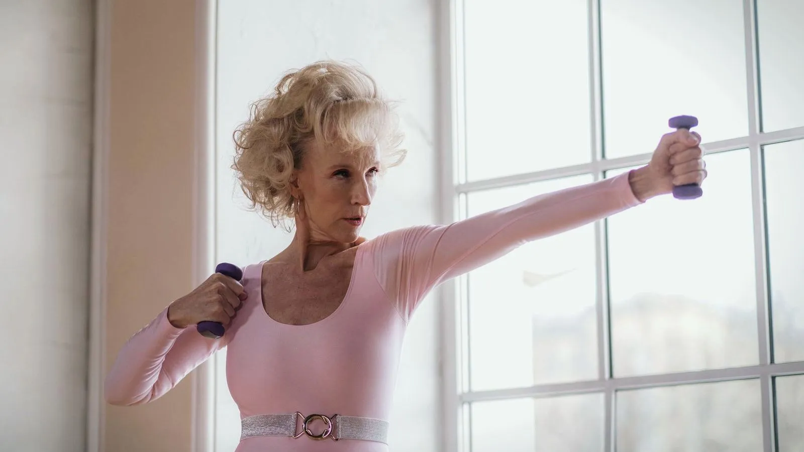 An older woman raising a pair of small dumbbells beside a tall window