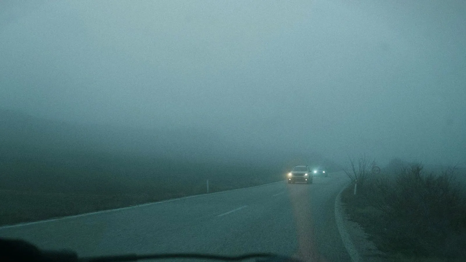 A car with its headlights on driving along a curving road in thick fog.