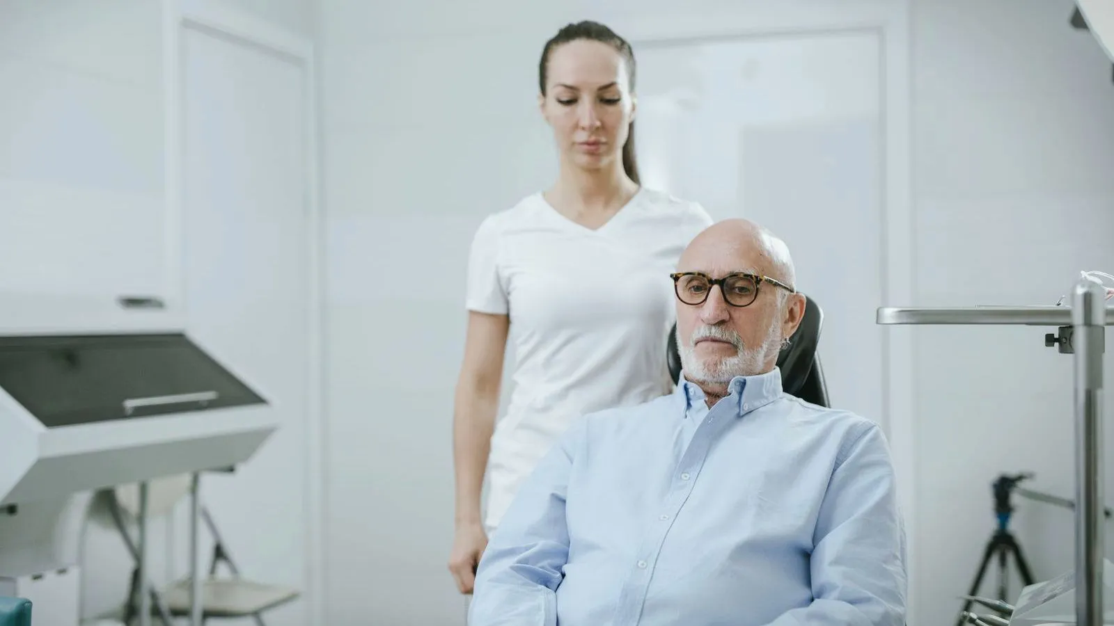 An older man with glasses and a white beard sits in a clinic chair while a woman in a white top stands behind him.