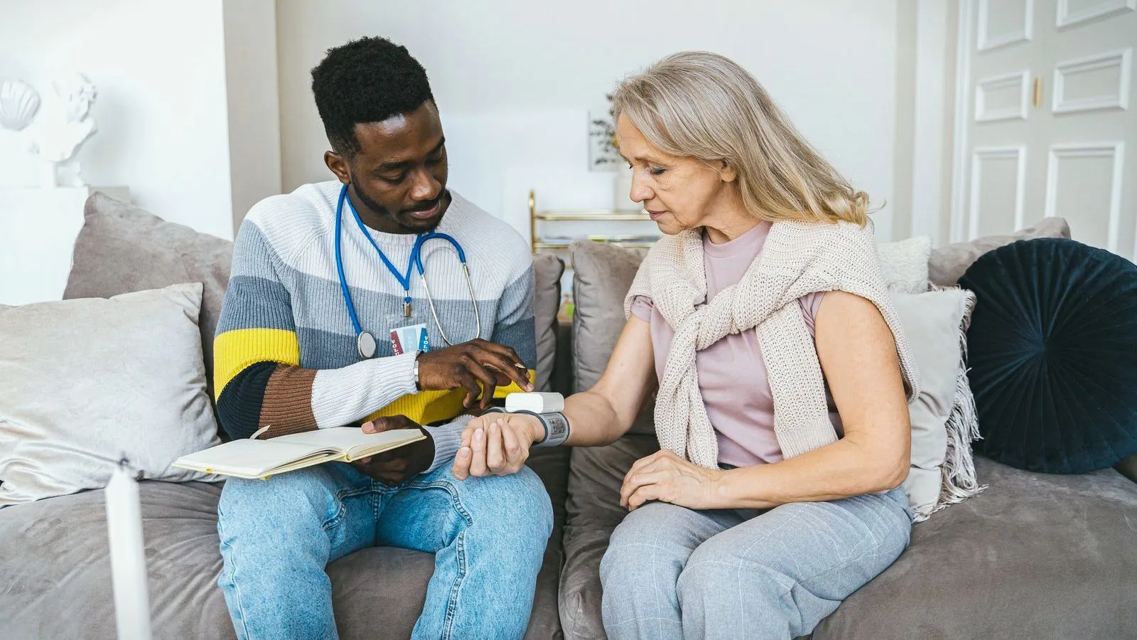 A clinician checking an older person's blood pressure as they sit together on a sofa.