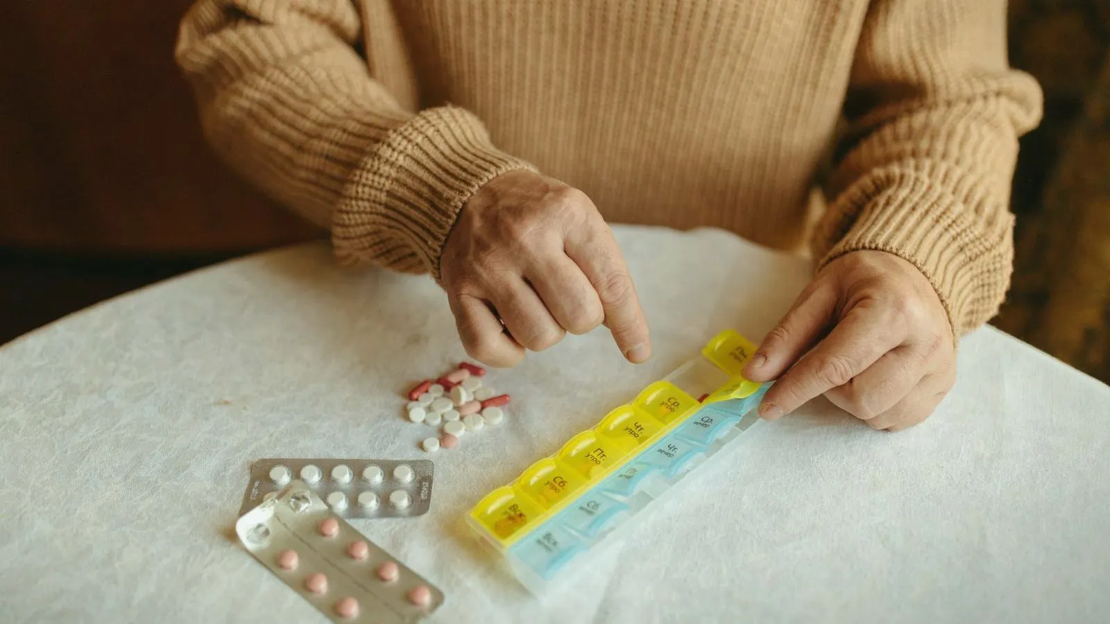 An older person's hands filling a weekly pill organizer beside loose tablets and blister packs
