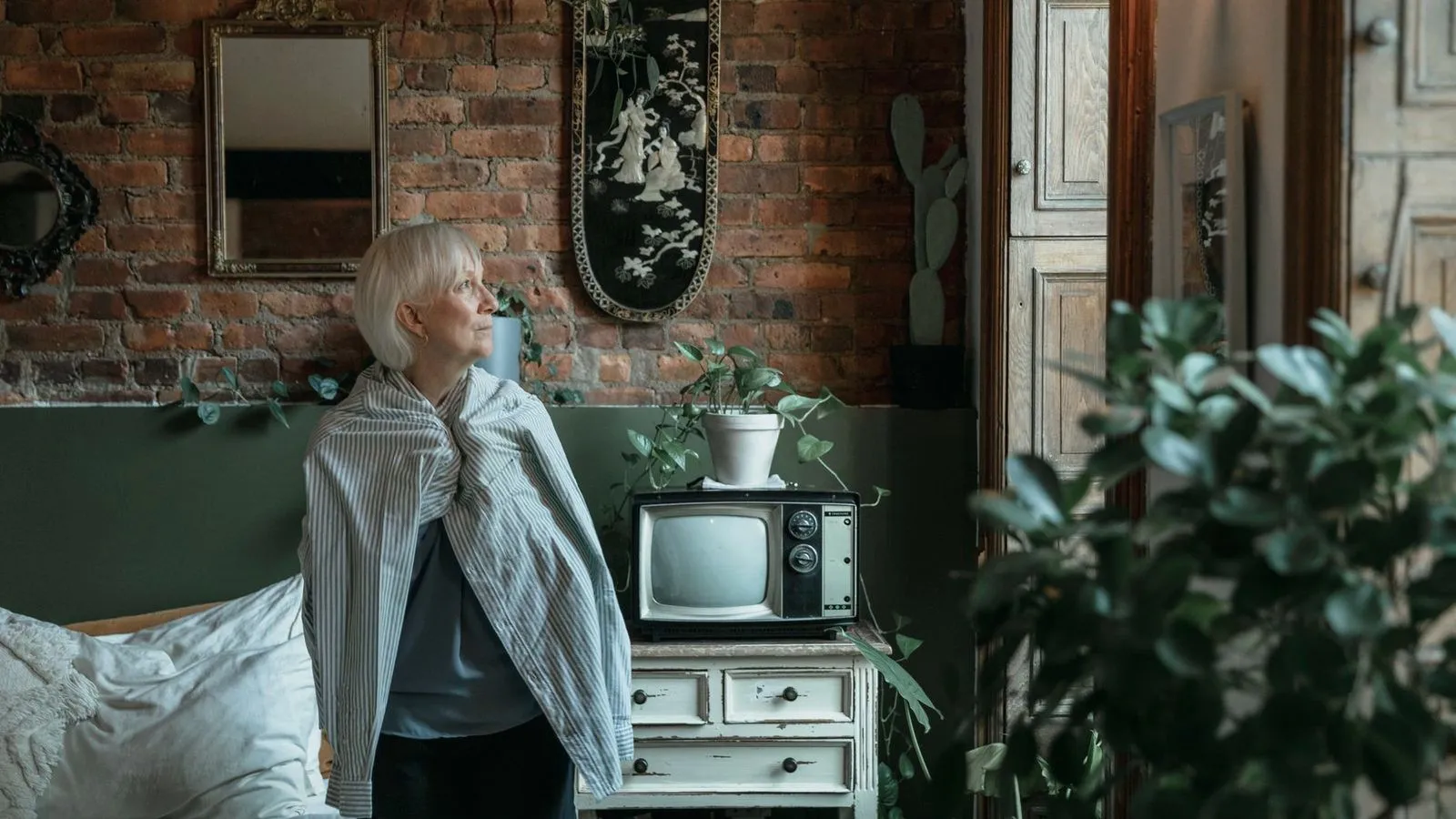 An older woman wrapped in a striped shirt looking away in a brick-walled room