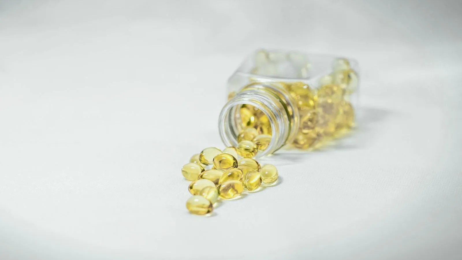 A tipped-over glass jar spilling yellow softgel capsules onto a white surface.