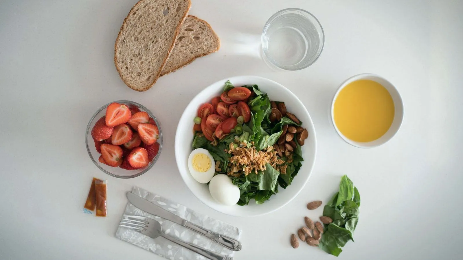 A meal seen from above: a salad with tomatoes, eggs and almonds, bread, strawberries and a glass of water