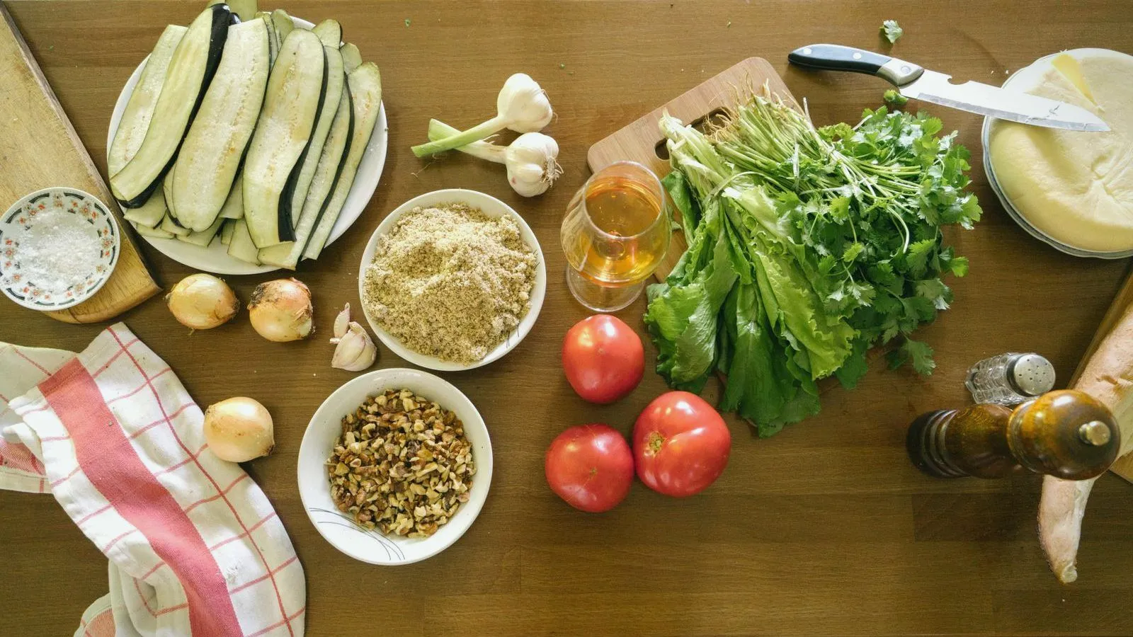 A wooden table seen from above with sliced eggplant, tomatoes, onions, garlic, leafy greens and bowls of grains and chopped nuts.