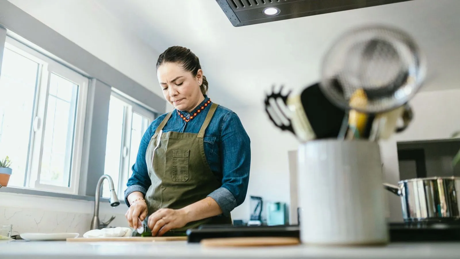 A woman in an apron chopping vegetables at a kitchen counter by a window