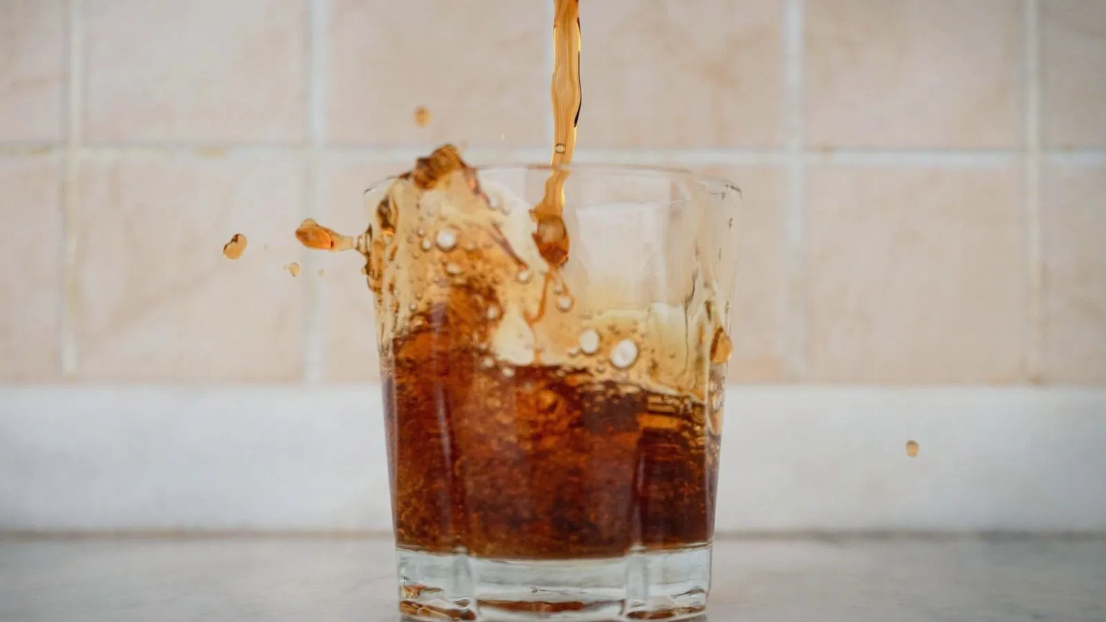 Dark cola pouring into a glass and splashing over the rim, in front of a tiled wall.