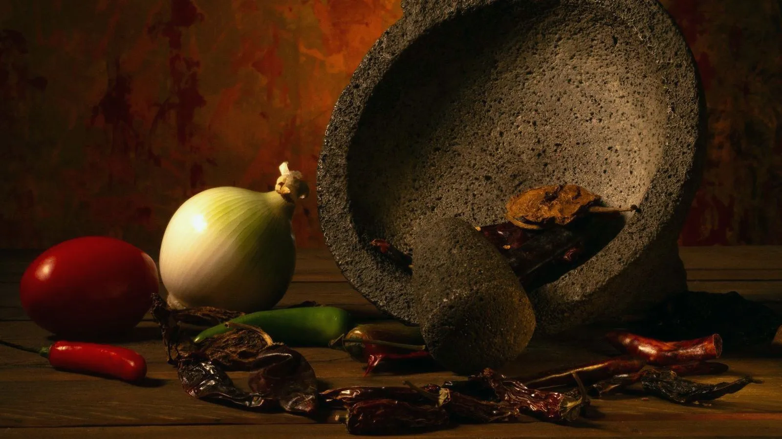 A stone mortar surrounded by dried chillies, tomato, onion and garlic in warm light