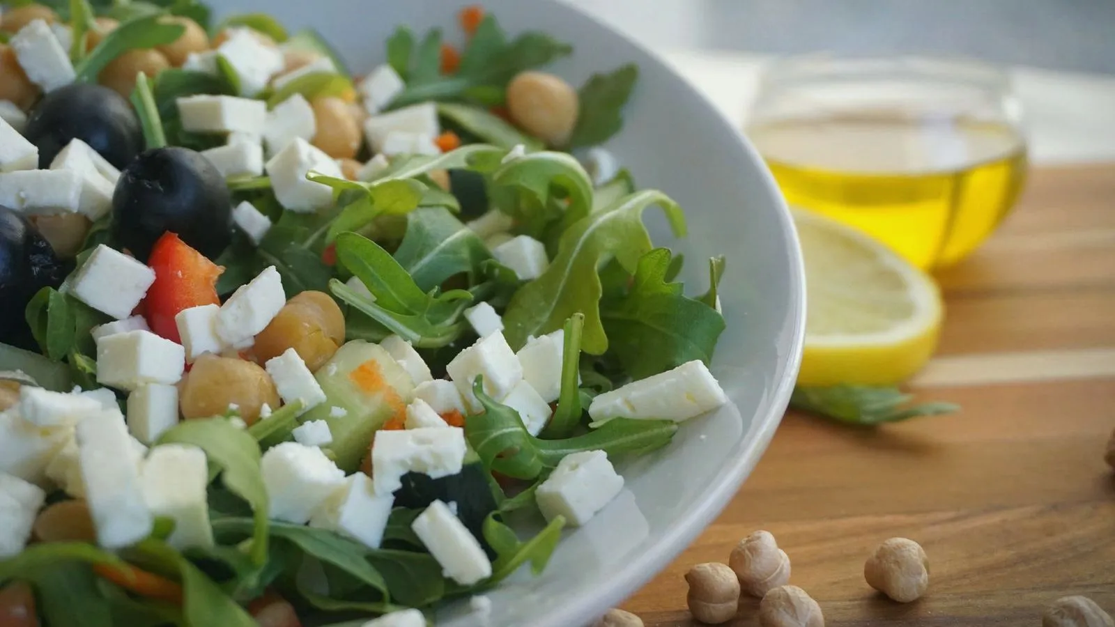 A bowl of salad with rocket, feta, olives, chickpeas and tomato, with olive oil and lemon behind it.