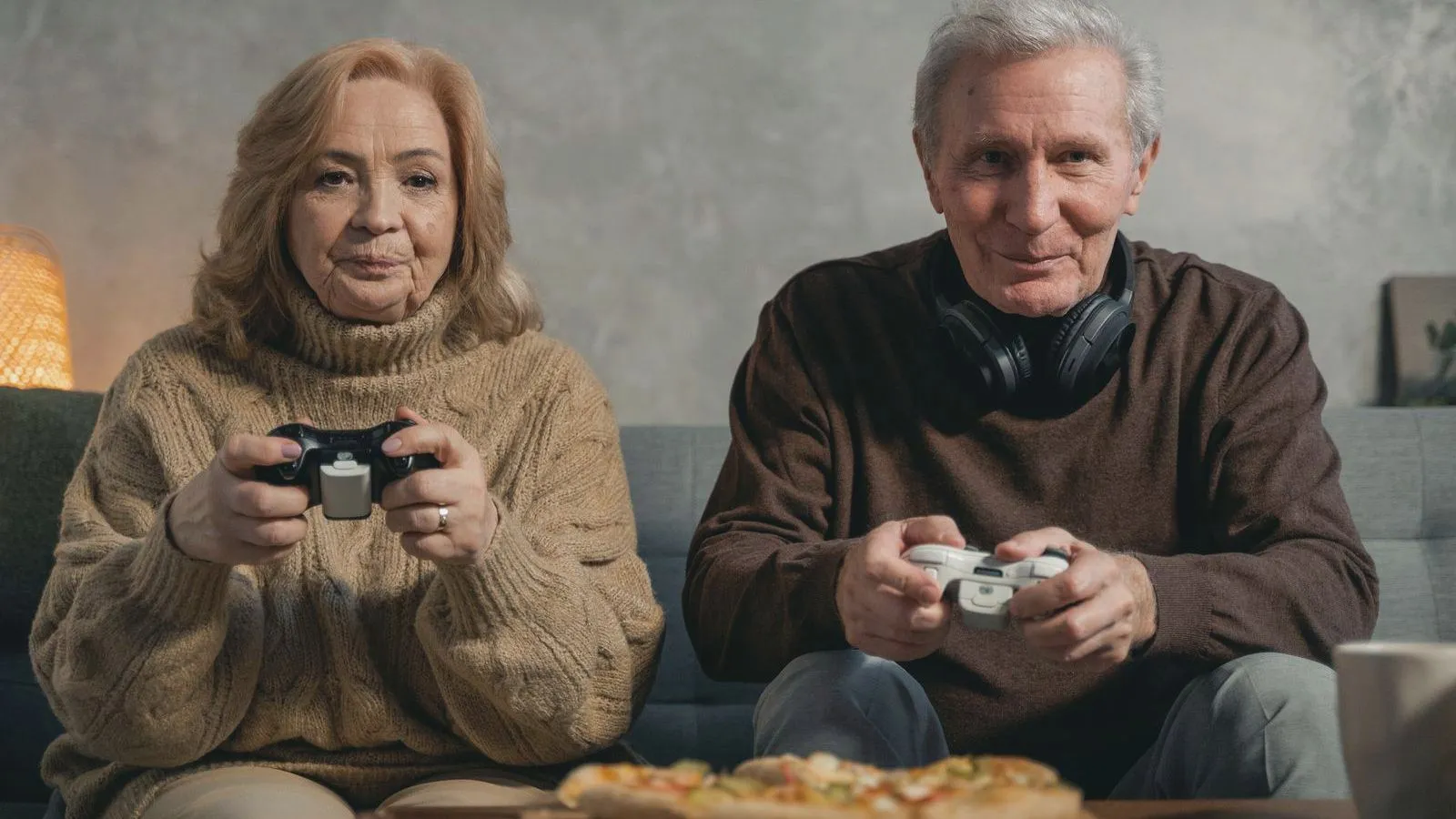 An older woman and man sitting on a sofa holding game controllers, a pizza on the table in front of them