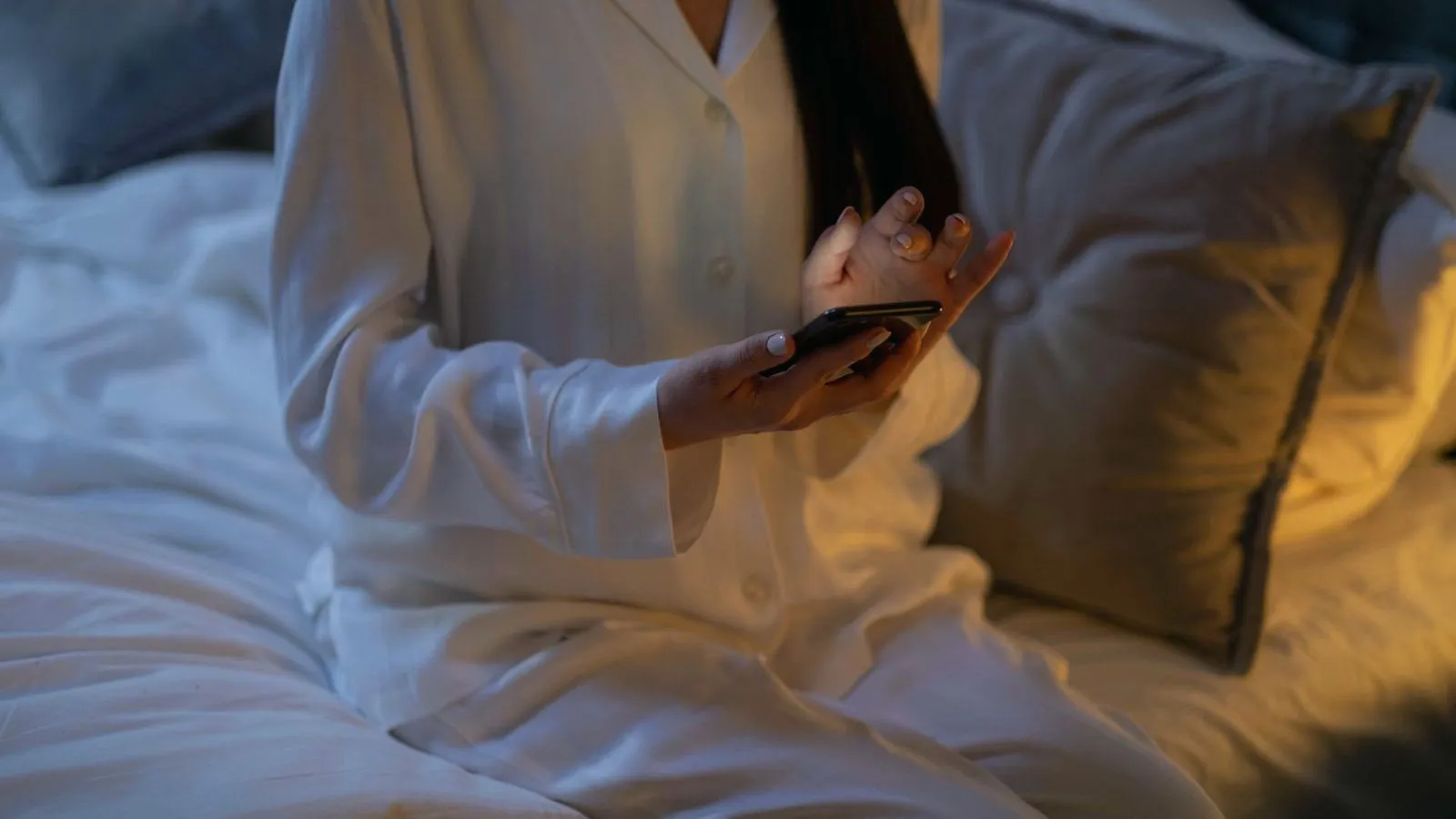 A person in white pajamas sitting on a bed and holding a smartphone, face not shown.