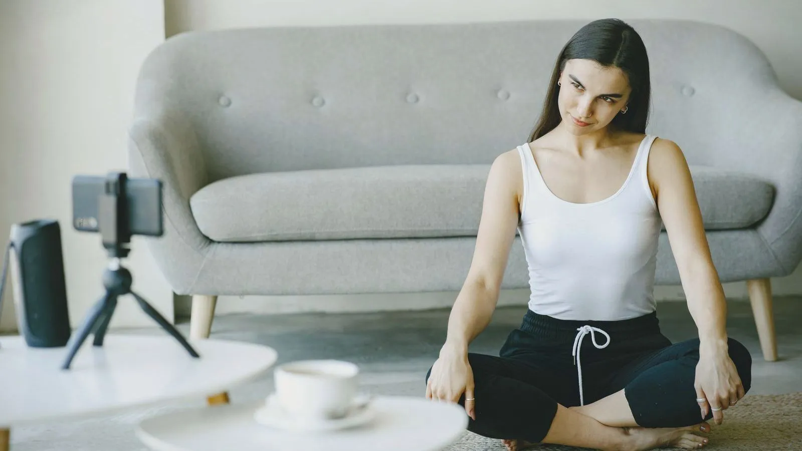 A woman sitting cross-legged on the floor by a sofa, following a session on a phone propped on a small tripod