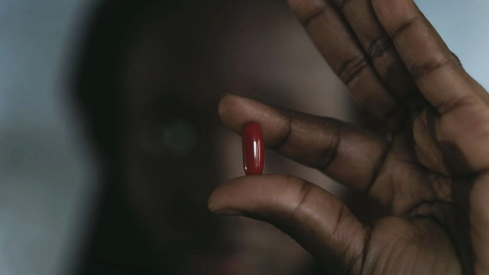 Fingers hold a single dark red capsule in front of a blurred background.