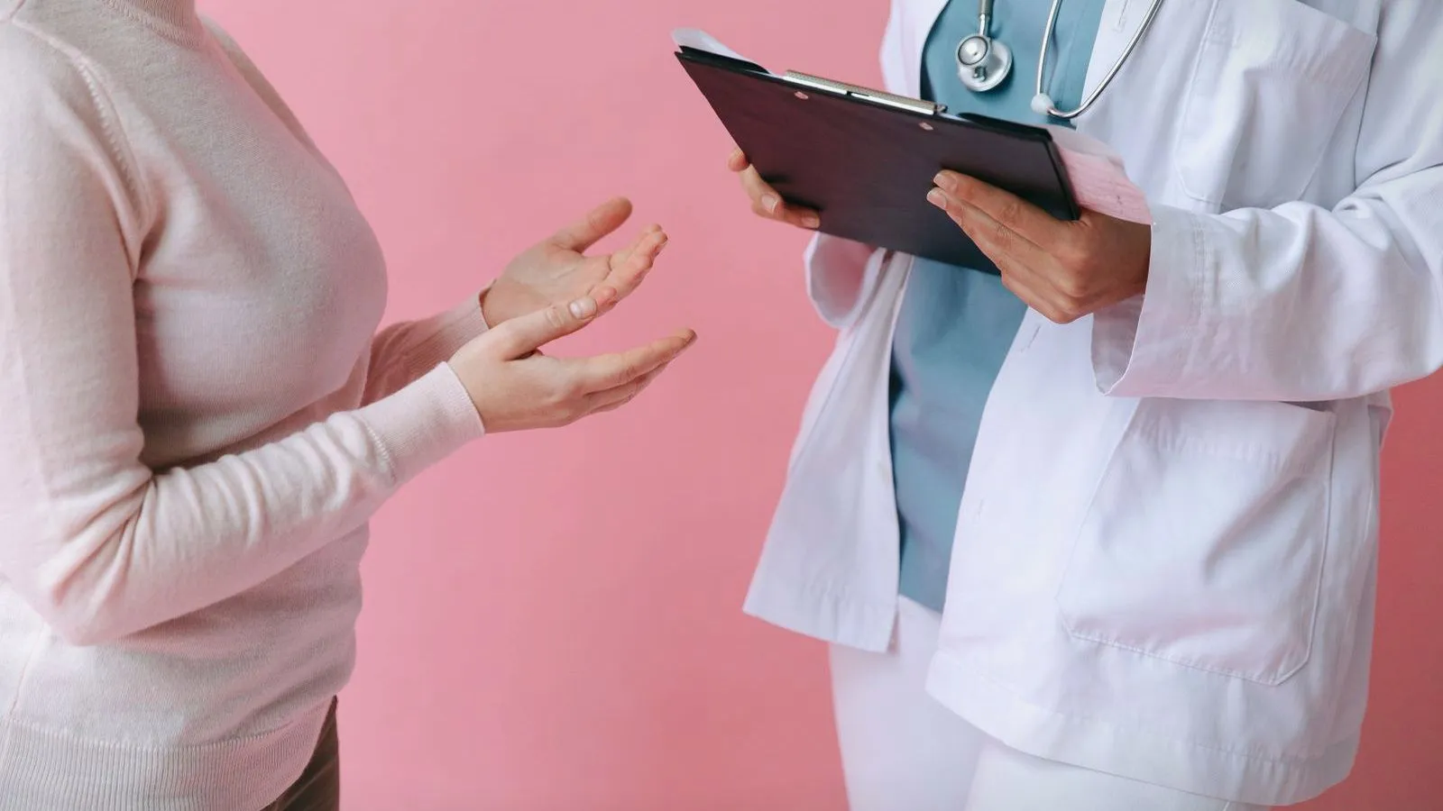 A person in a pink sweater gestures with open hands while talking to a clinician in a white coat who holds a clipboard; neither face is shown.