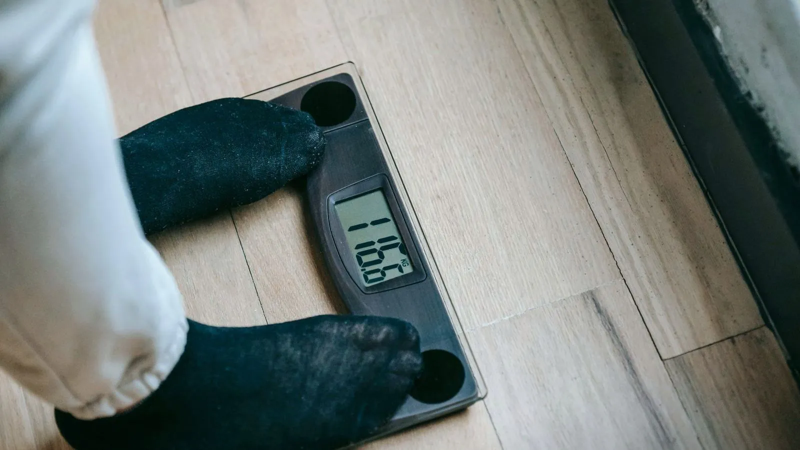 A pair of feet in dark socks standing on a digital bathroom scale on a wooden floor