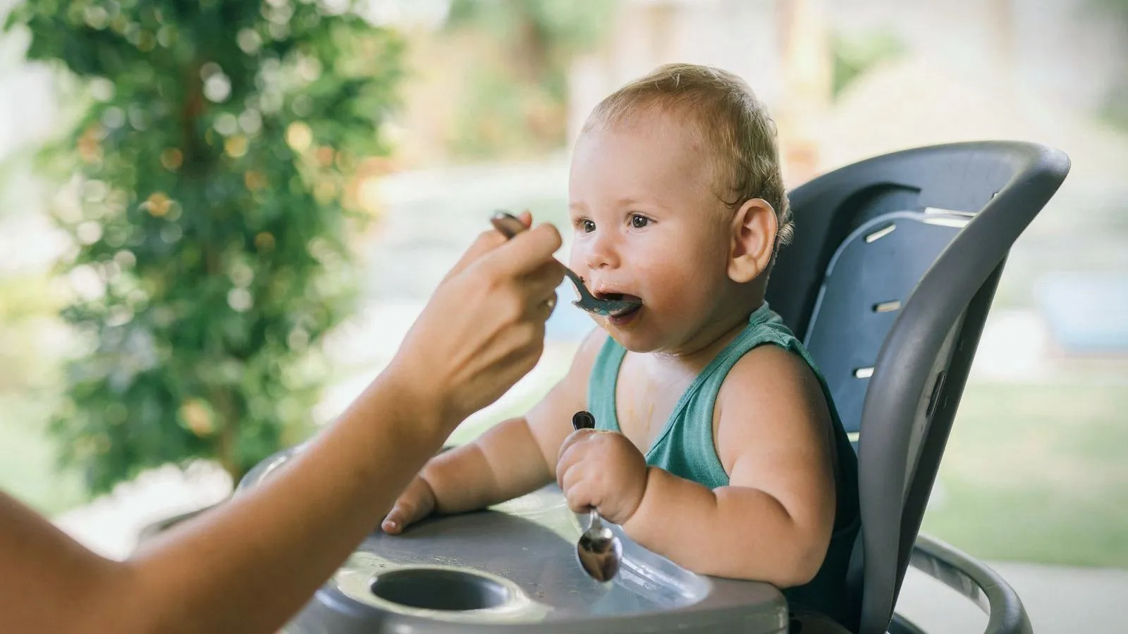 A baby in a high chair being fed from a spoon while holding a spoon of their own