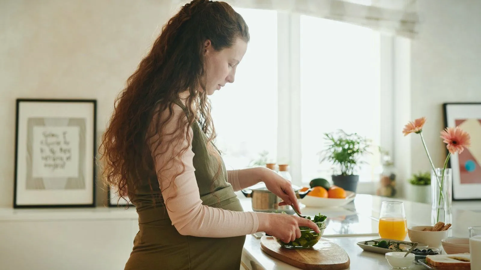 A pregnant woman preparing a salad at a sunny kitchen counter