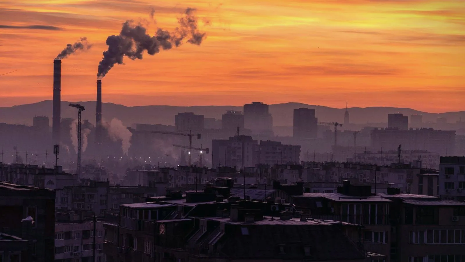 Smokestacks releasing smoke over a hazy city skyline at sunset