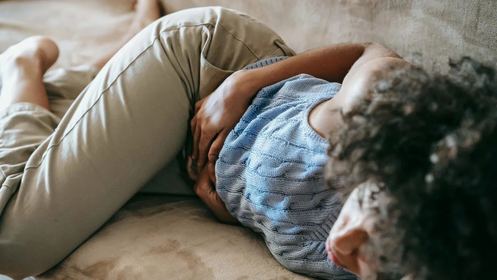 A person curled on their side on a sofa with both hands pressed to their abdomen