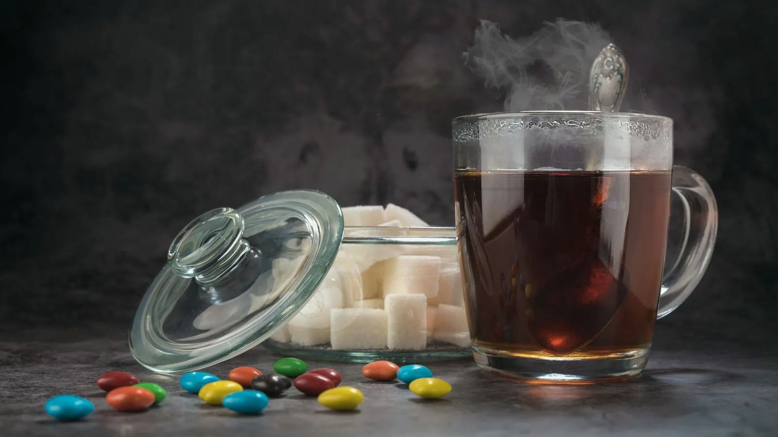 A glass mug of tea beside sugar cubes and candy-coated sweets on a dark surface