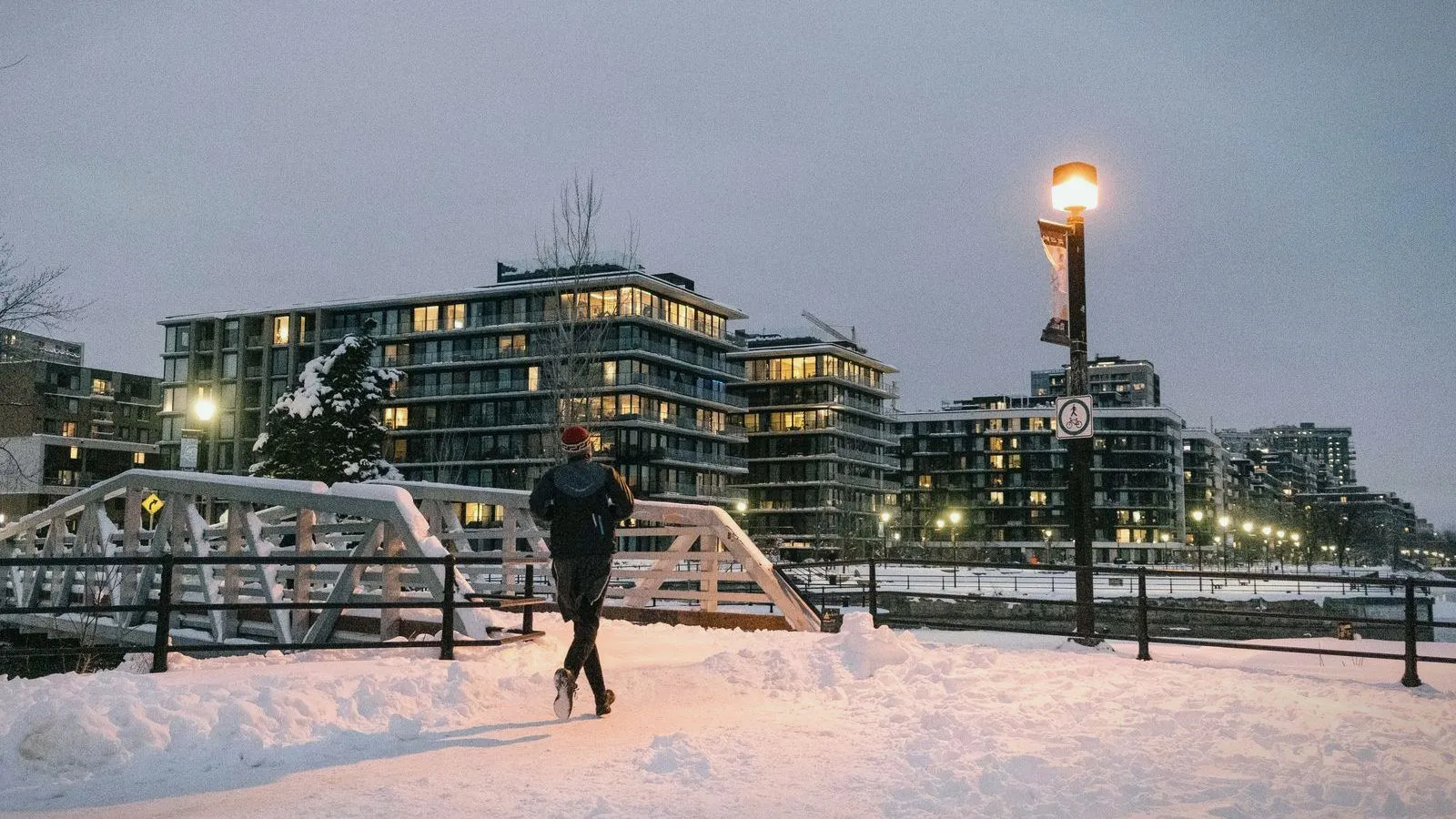 A runner on a snow-covered path at dusk, with a lit street lamp and apartment buildings behind.