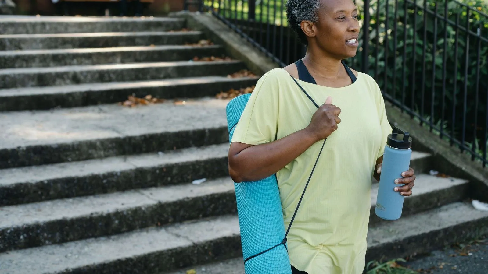 A person in a yellow top walks past concrete park steps with a rolled exercise mat on one shoulder and a water bottle.