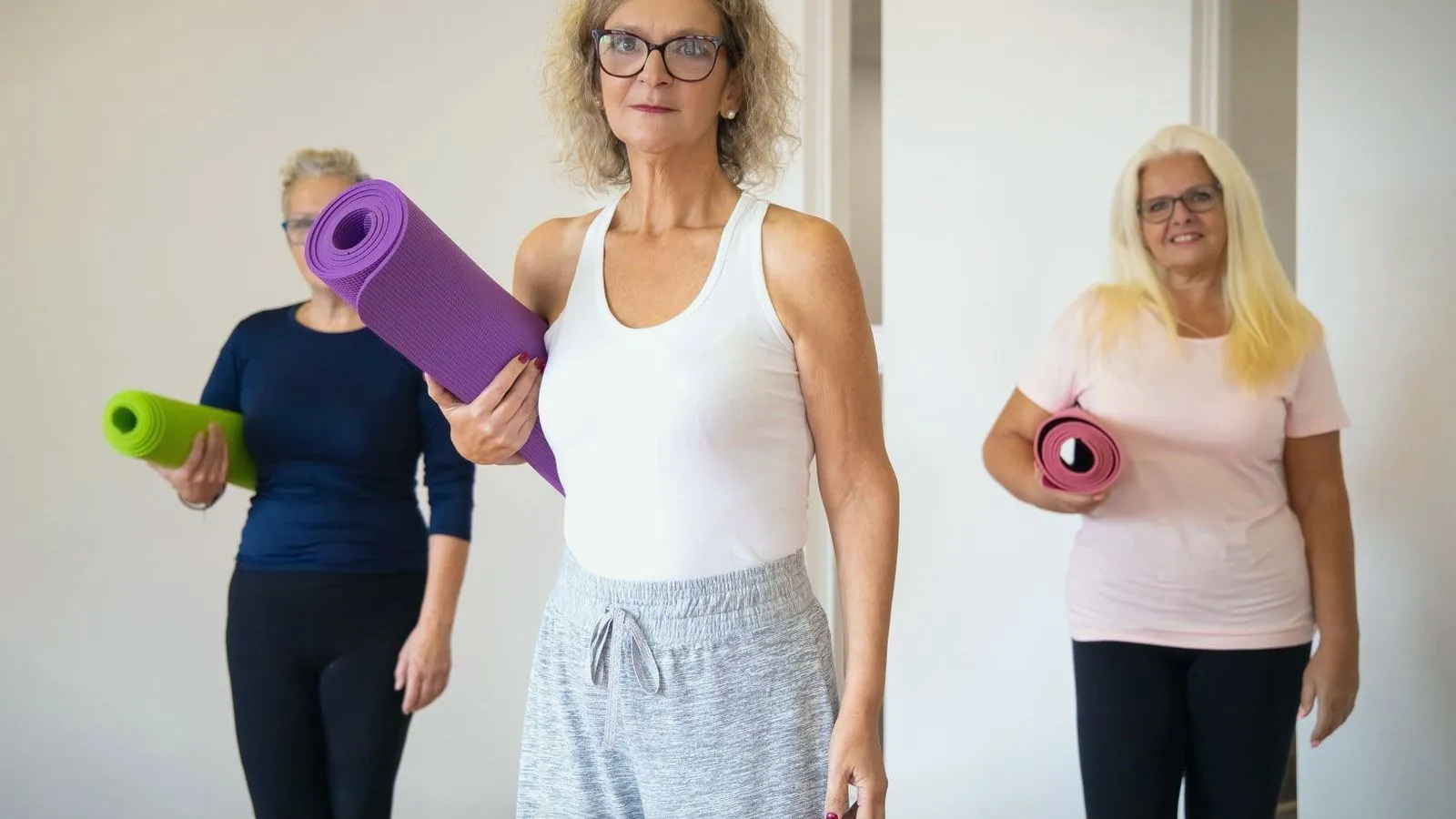 Three older women holding rolled exercise mats in a bright studio.