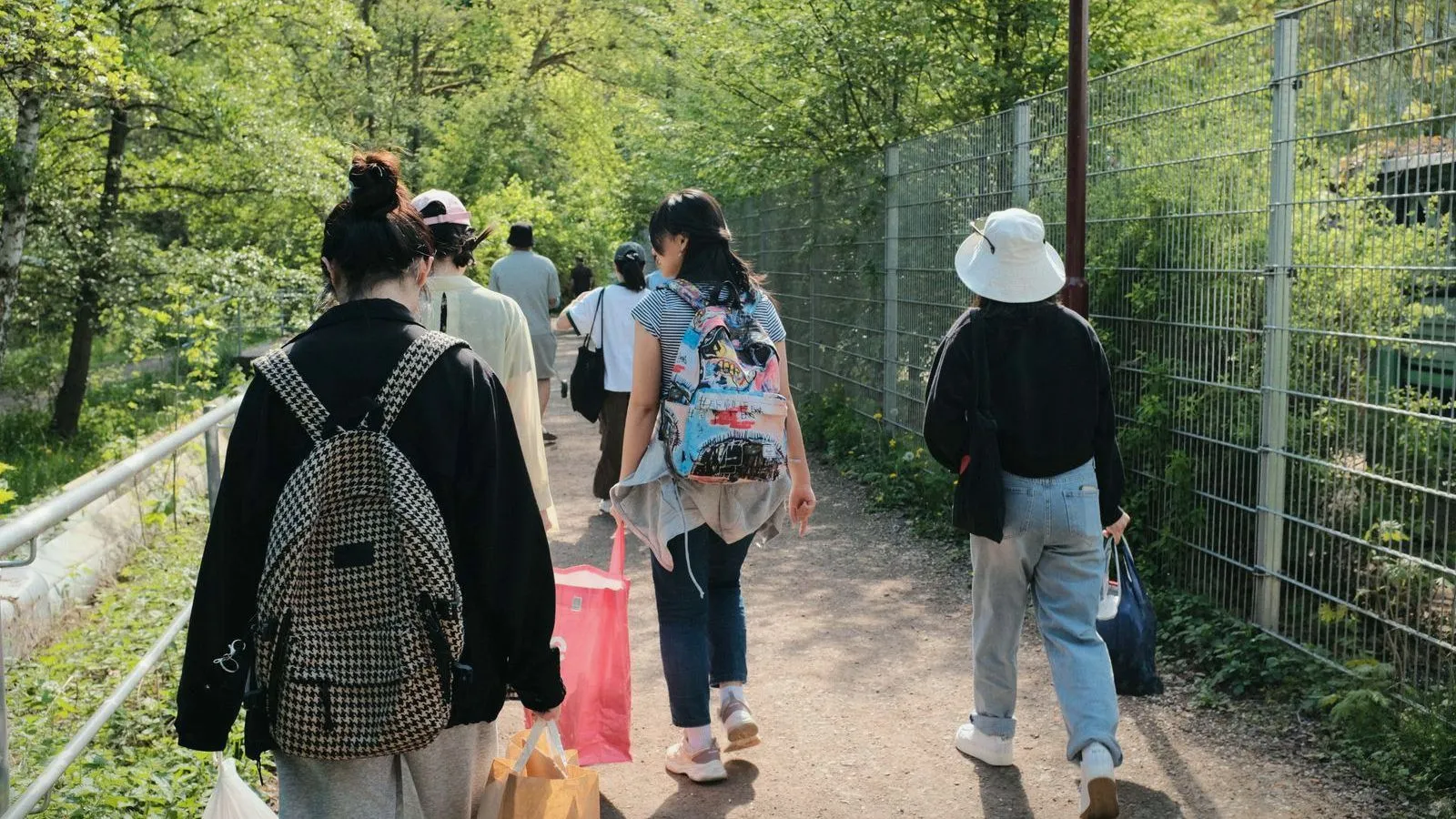 Several people with backpacks and bags walking away along a path beside a wire fence, under trees.