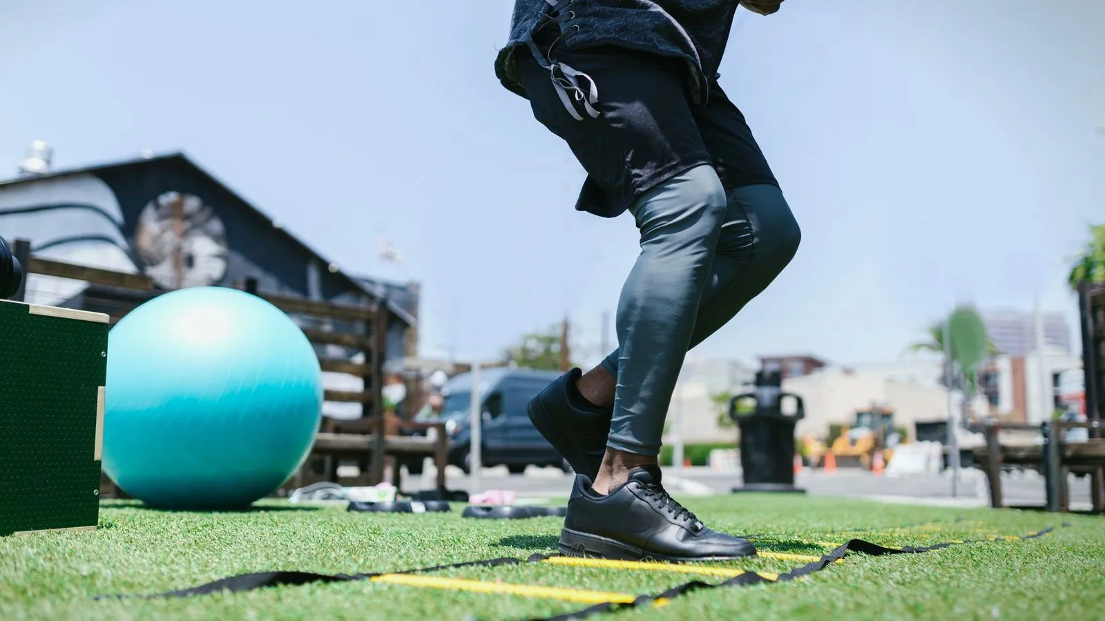 Someone running through an agility ladder on outdoor turf beside an exercise ball