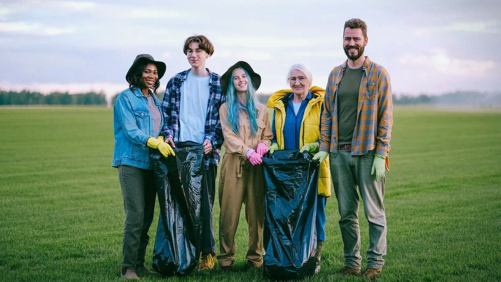 Five volunteers in gloves holding rubbish bags on a wide grassy field