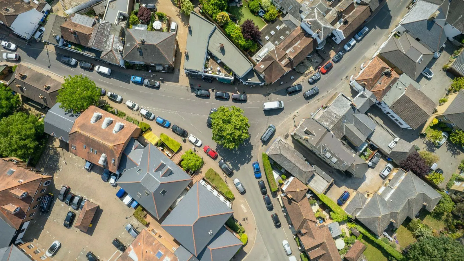 Aerial view of a town junction with tiled roofs, gardens, trees and cars parked along curving streets.