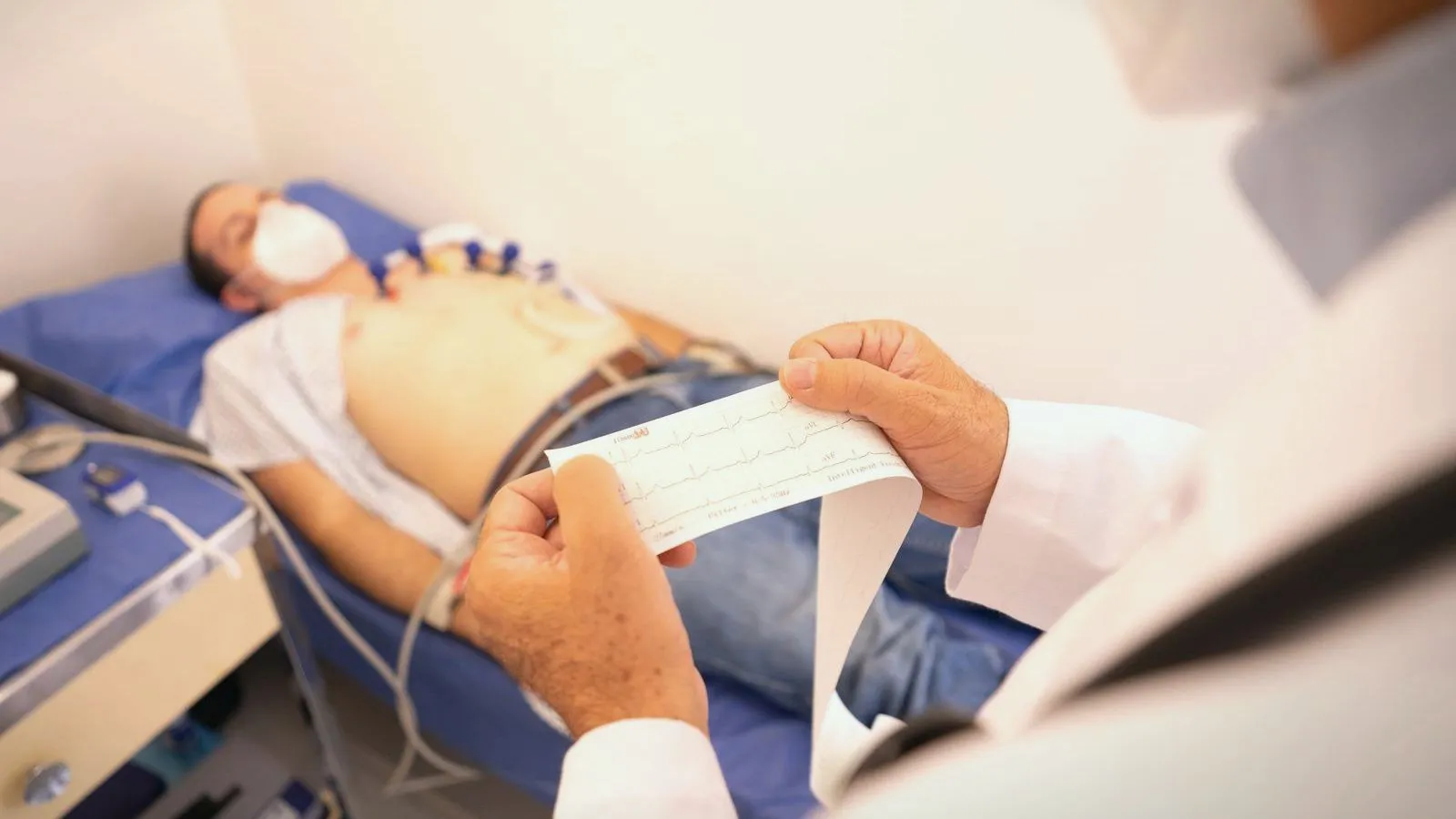 A patient lying with heart monitor leads on the chest while a clinician reads a printed heart tracing