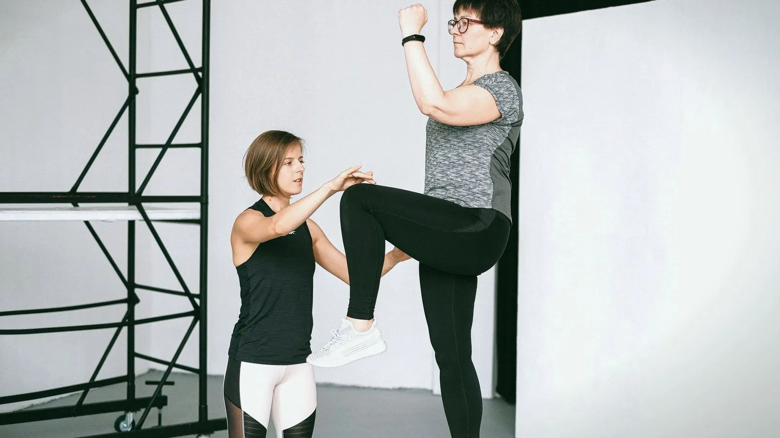 A trainer steadies a woman as she balances on one leg with a knee raised in a bright studio.