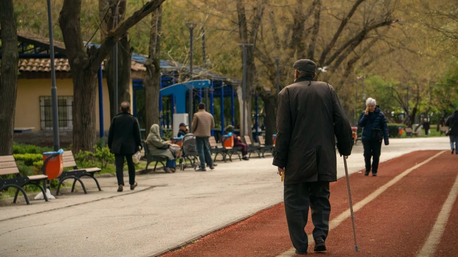 An older man in a dark coat walking with a cane along a park path, other people behind him