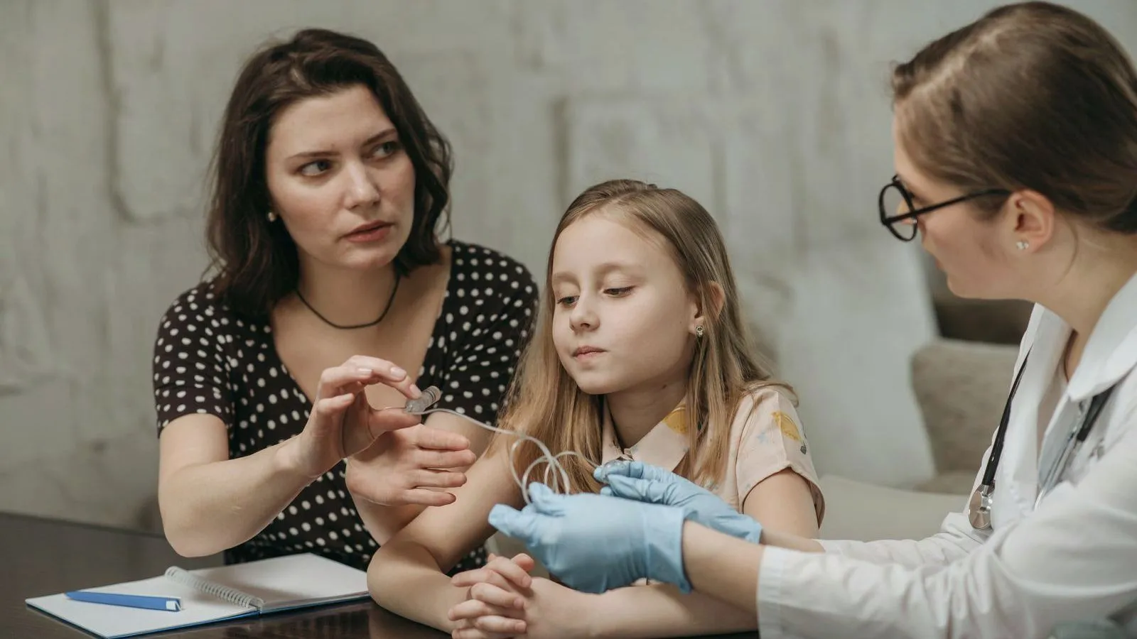 A clinician in blue gloves attends to a child's arm while a parent sits close beside them at a table