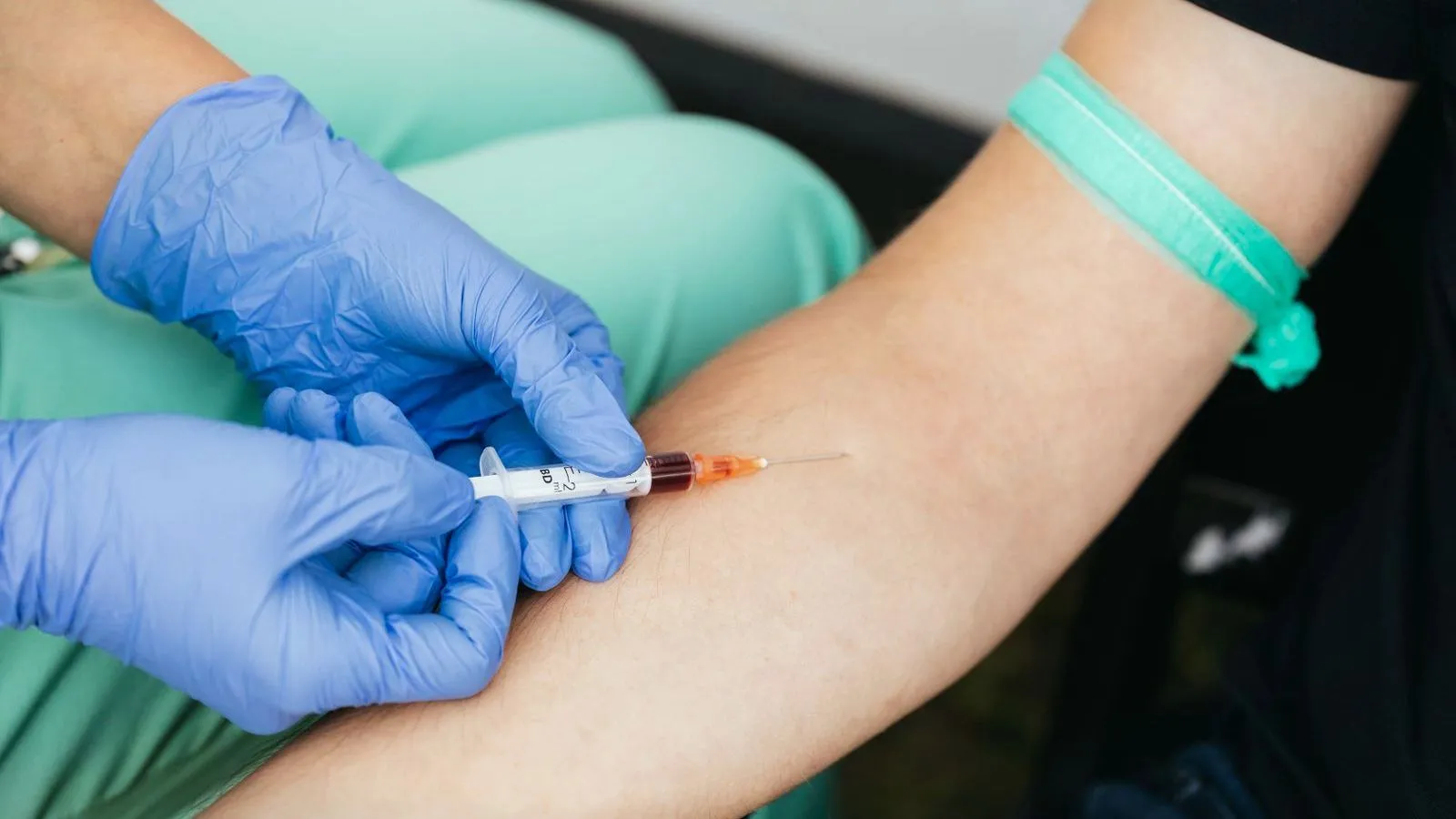Hands in blue gloves draw blood with a syringe from the inside of an arm that has a teal tourniquet above the elbow.