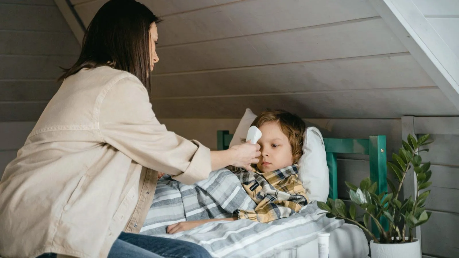 A parent checking a sick child's temperature with a forehead thermometer as the child lies in bed.