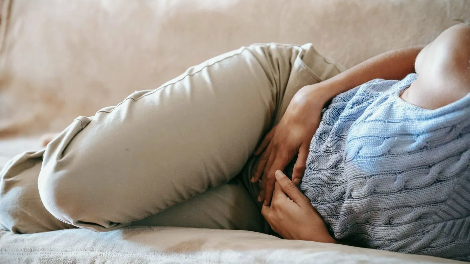 A person lies curled on a sofa with both hands pressed to the abdomen, face out of frame.