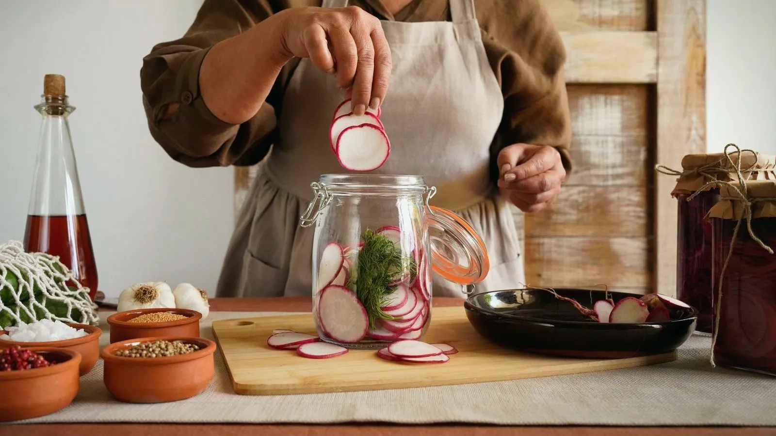 A person in an apron adding sliced radishes to a glass jar on a kitchen counter.