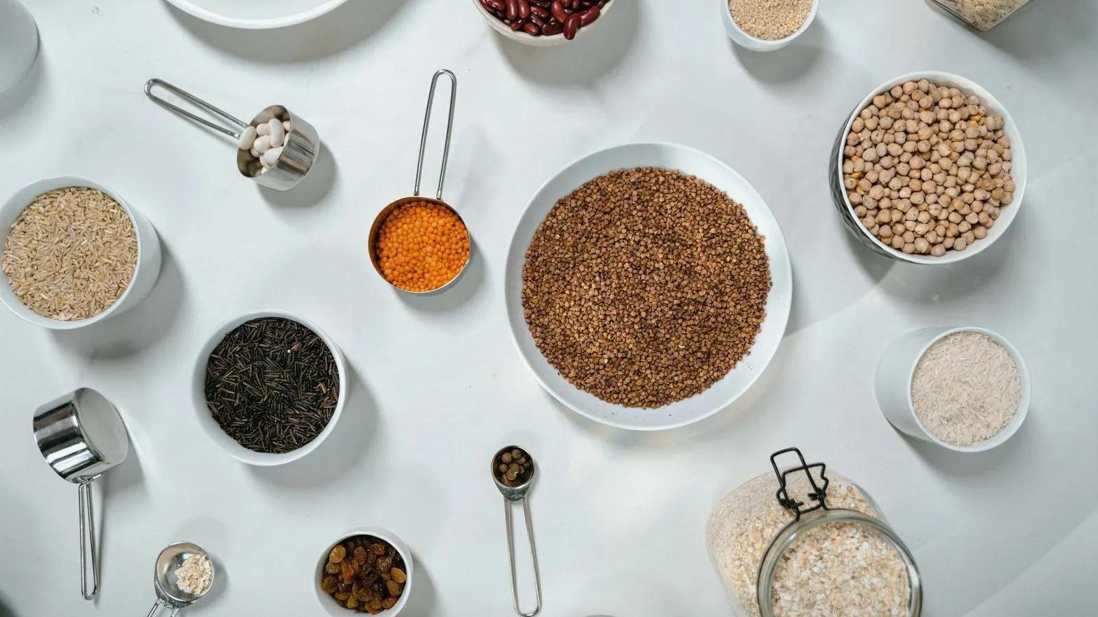 Bowls and measuring cups of grains, lentils, chickpeas and wild rice arranged on a white surface, seen from above.