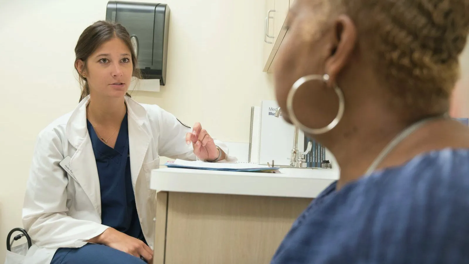 A clinician in a white coat talking with a patient across a consulting room.