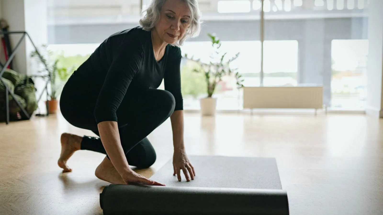 An older person with grey hair, dressed in black, kneels barefoot to unroll an exercise mat in a bright studio.
