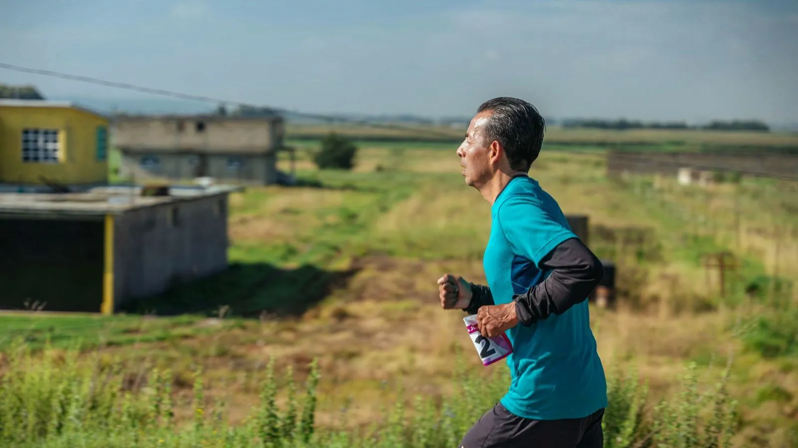 An older man in a teal running shirt jogging along a rural track, with fields and low buildings behind him