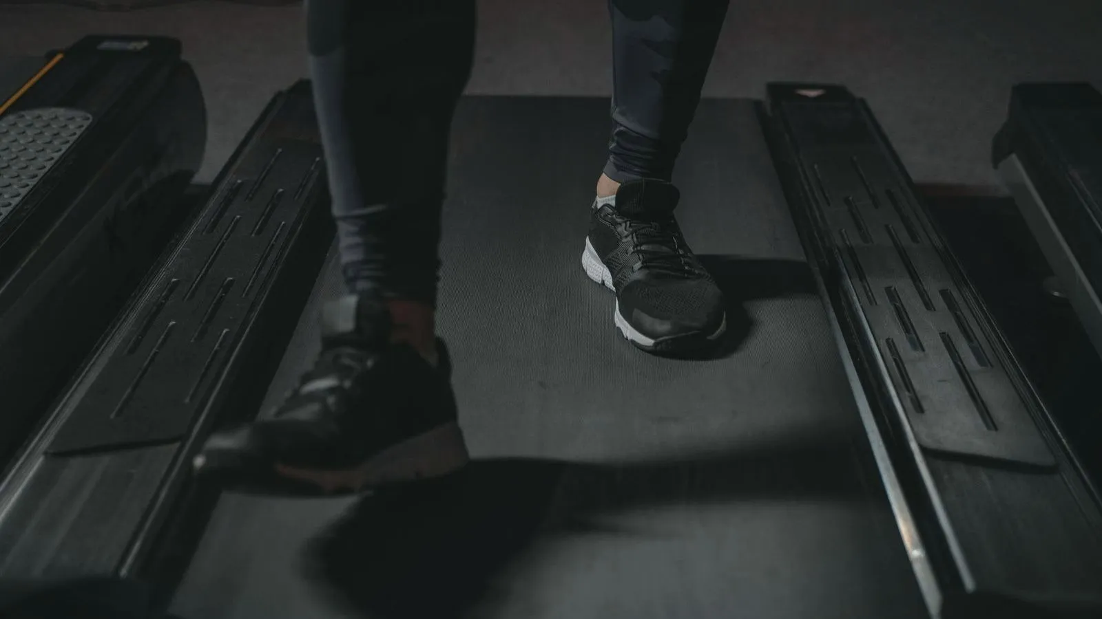 The lower legs and black trainers of a person walking on a dark treadmill belt, seen from above.