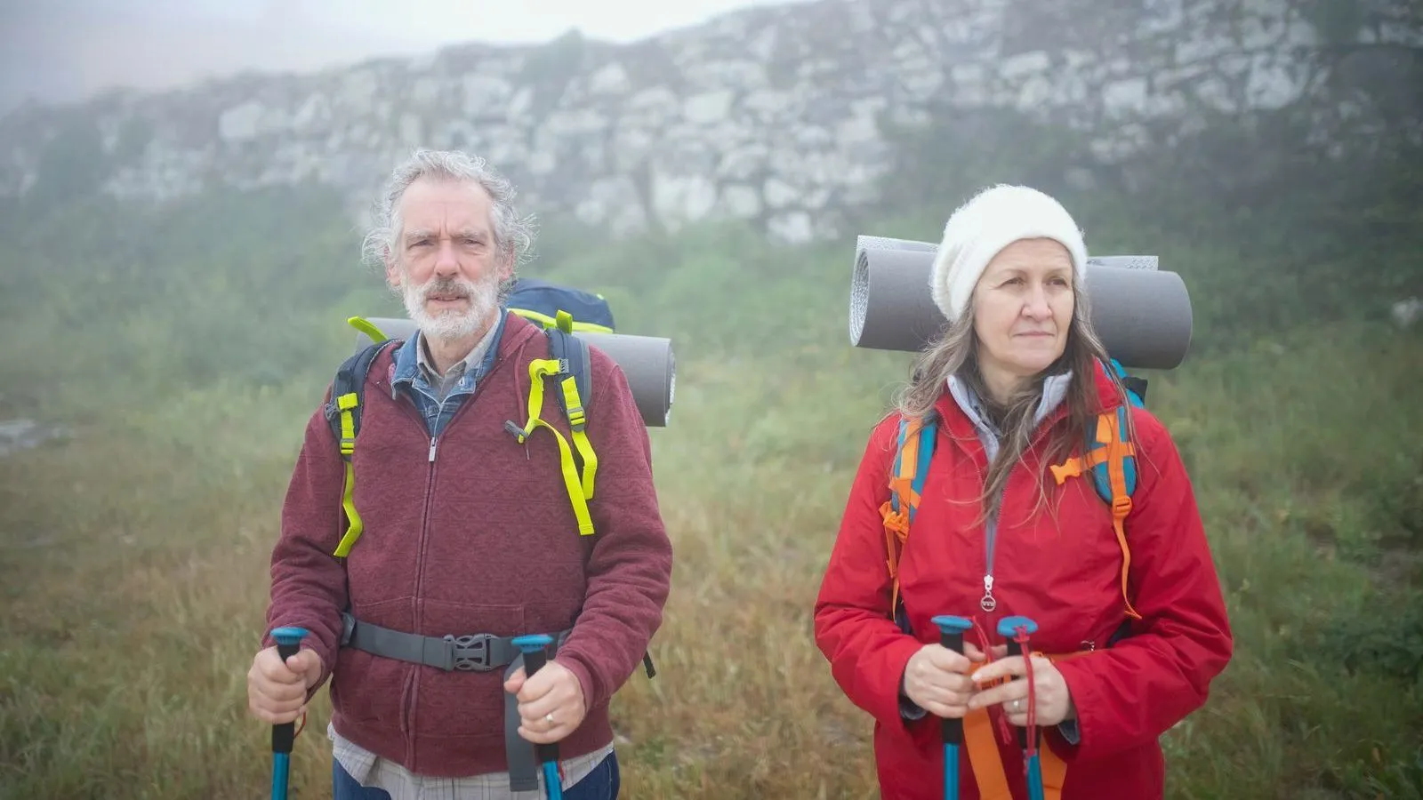 Two older hikers with backpacks and walking poles standing outdoors on a misty day.