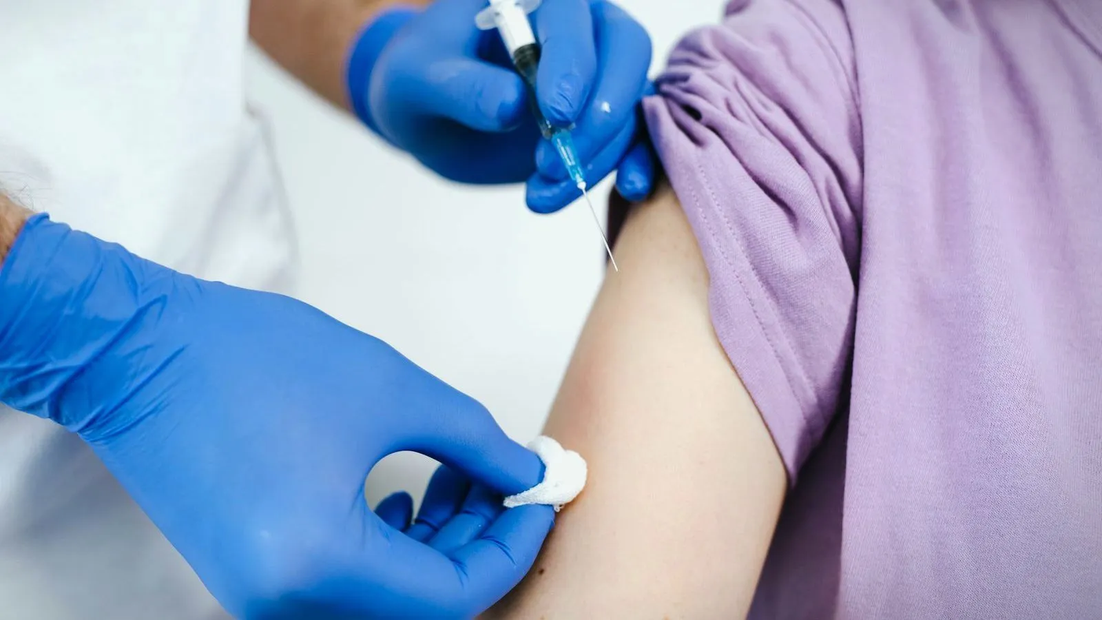 Hands in blue gloves hold a syringe and press a cotton swab to a person's upper arm below a rolled-up purple sleeve.