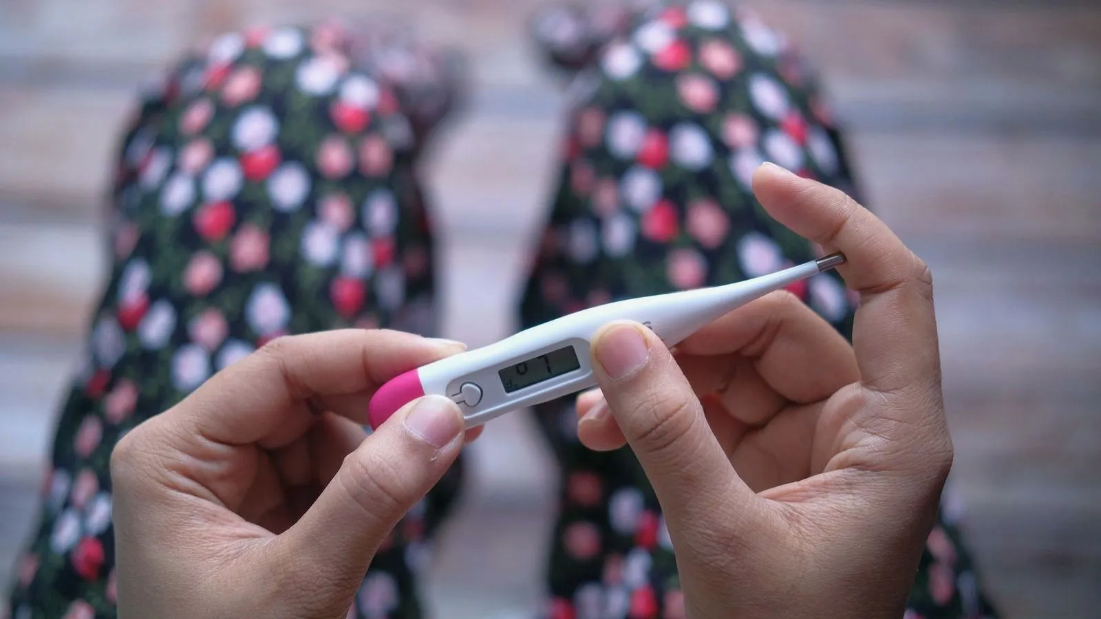 Two hands hold a digital thermometer with a pink tip in front of floral-patterned fabric.